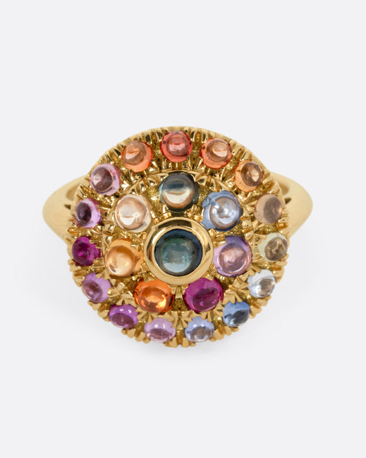 A yellow gold circular statement ring with multicolored round sapphire cabochons winding its face, shown from the front.