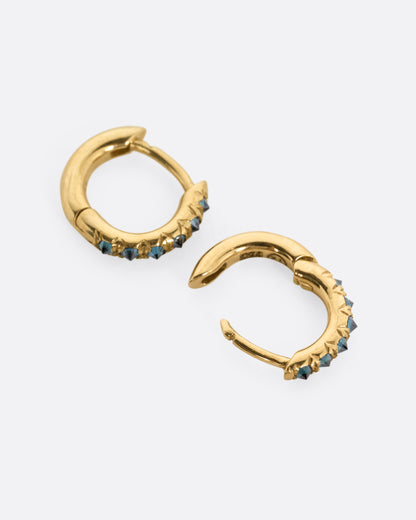 A pair of yellow gold huggie hoop earrings with reverse set blue diamonds, shown from the side with one earring open.
