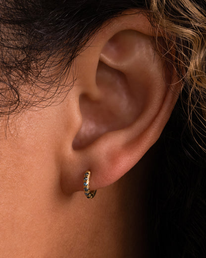 A yellow gold huggie hoop earring with reverse set blue diamonds, shown on a person.