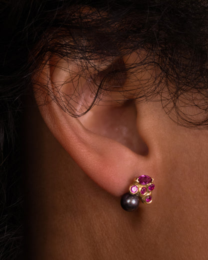 A yellow gold stud earring with rubies and a black pearl. Shown on a persn