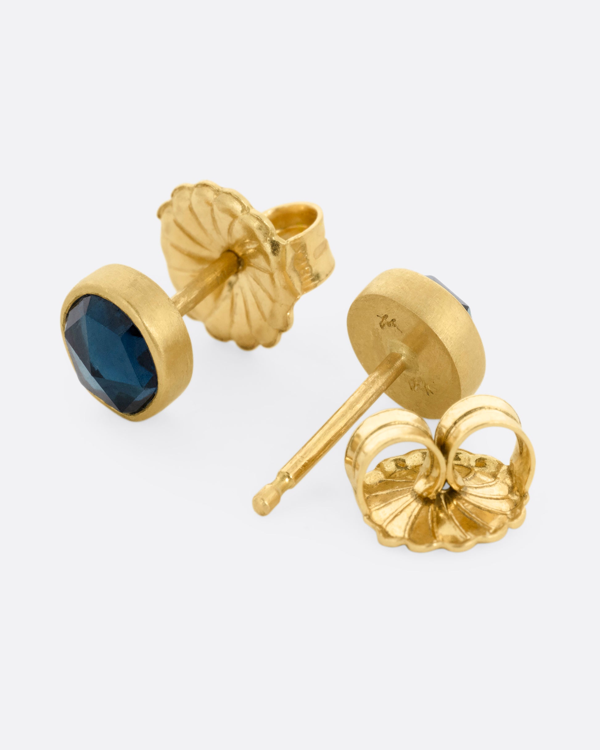 A pair of yellow gold stud earrings with blue green tourmalines. Both earrings are shown from the side. One earring is facing the front, the other is facing the back, open.