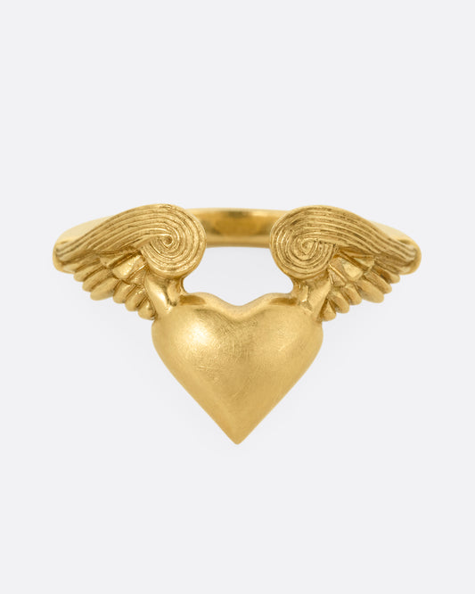 A yellow gold ring with a heart with wings on it. Shown from the front.