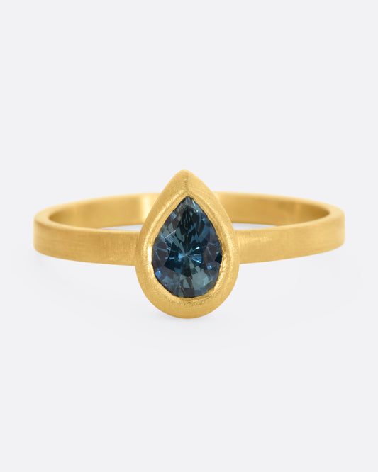 A yellow gold ring with a teal teardrop shaped sapphire. Shown from the front.