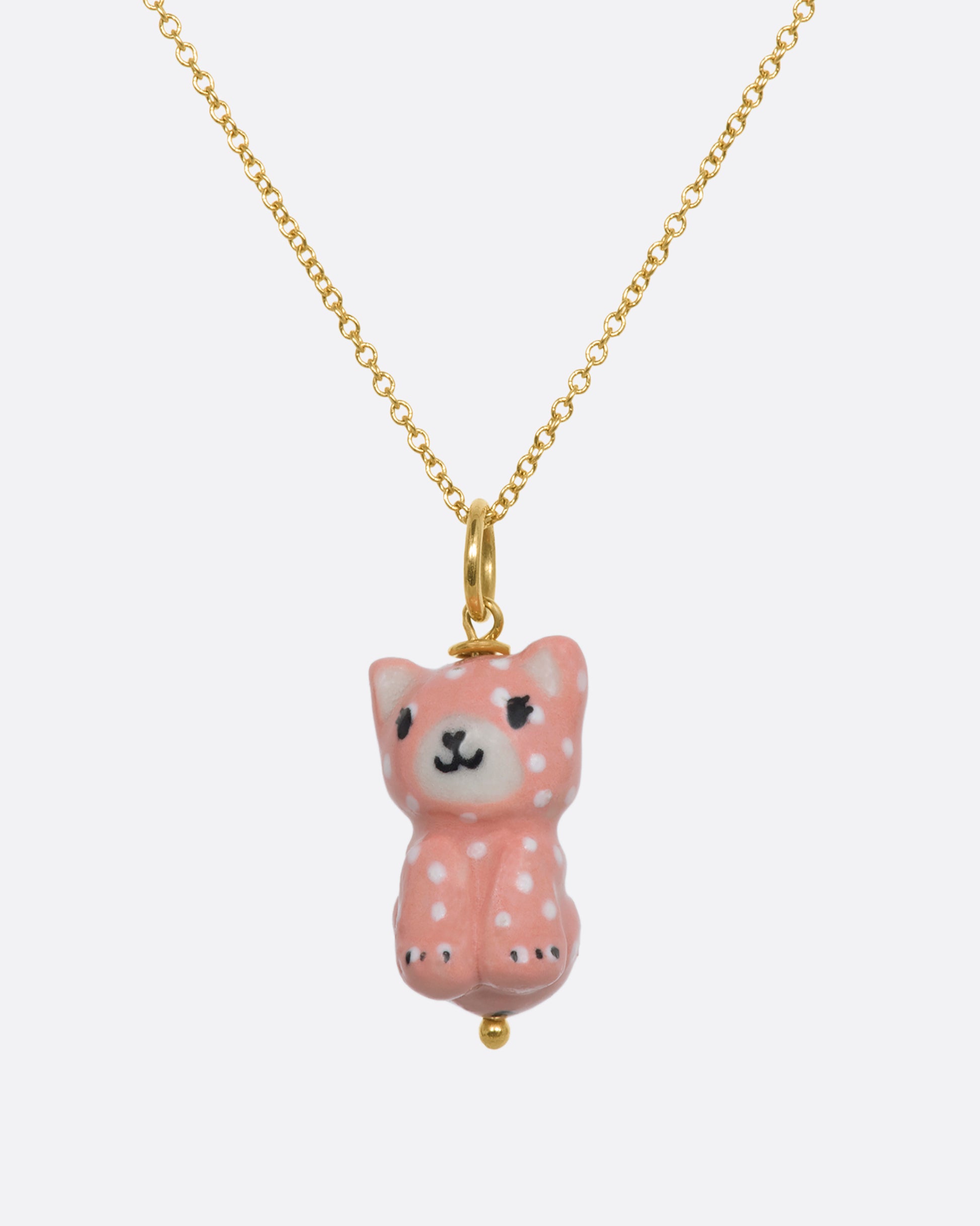 A three dimensional porcelain pink cat charm with white spots. Hanging from a yellow gold chain.