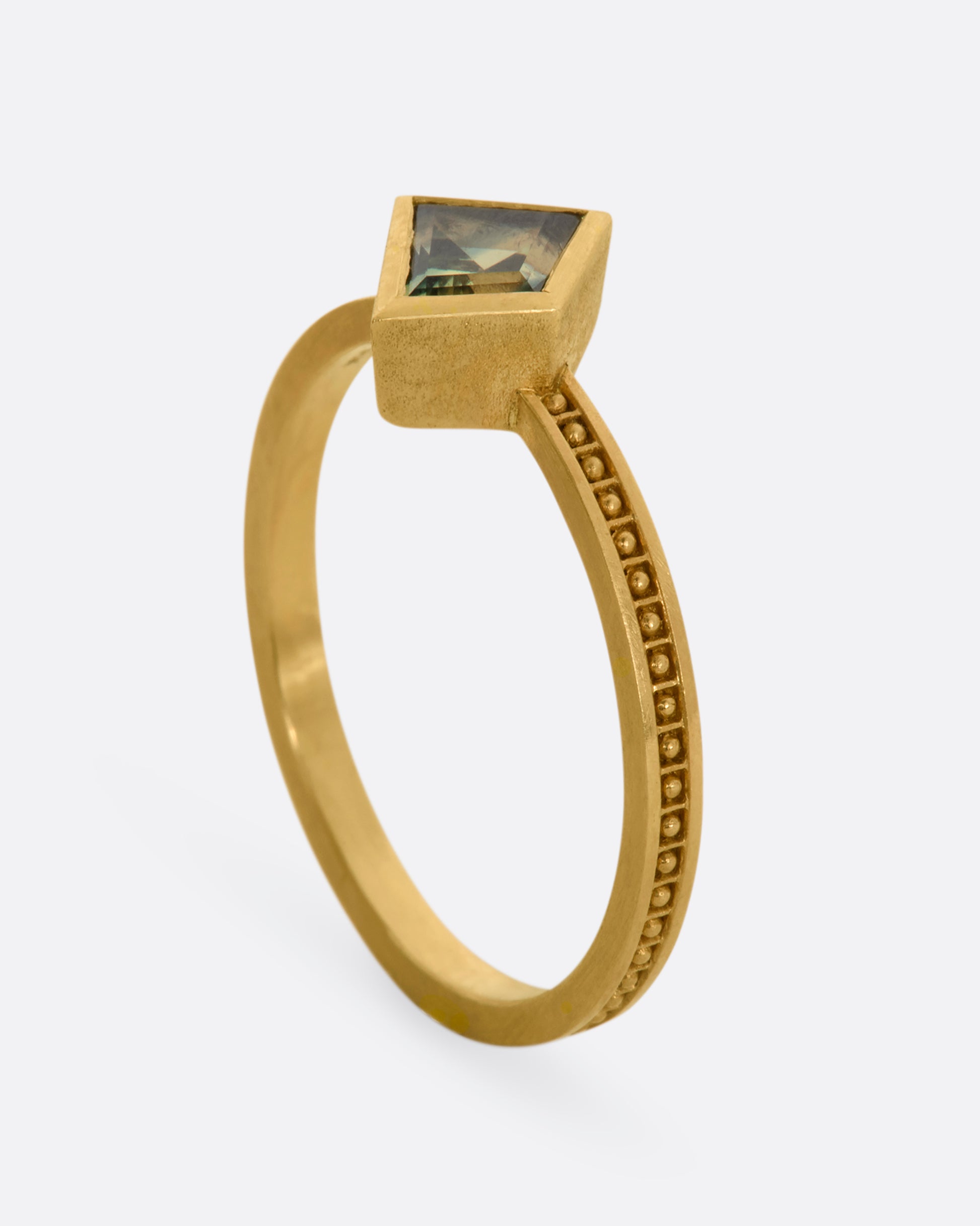 A yellow gold ring with a trapezoid green sapphire and raised dots in square shaped holes around the band. View standing.