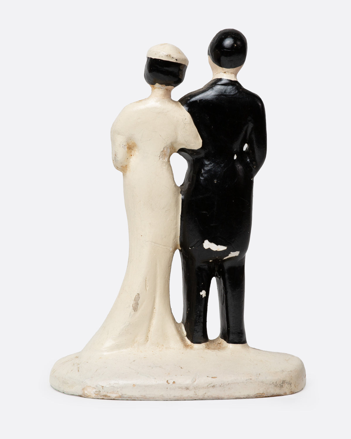 A ceramic wedding cake topper with a groom in a black tuxedo and a bride in a cream gown with pink floral details, shown from the back.
