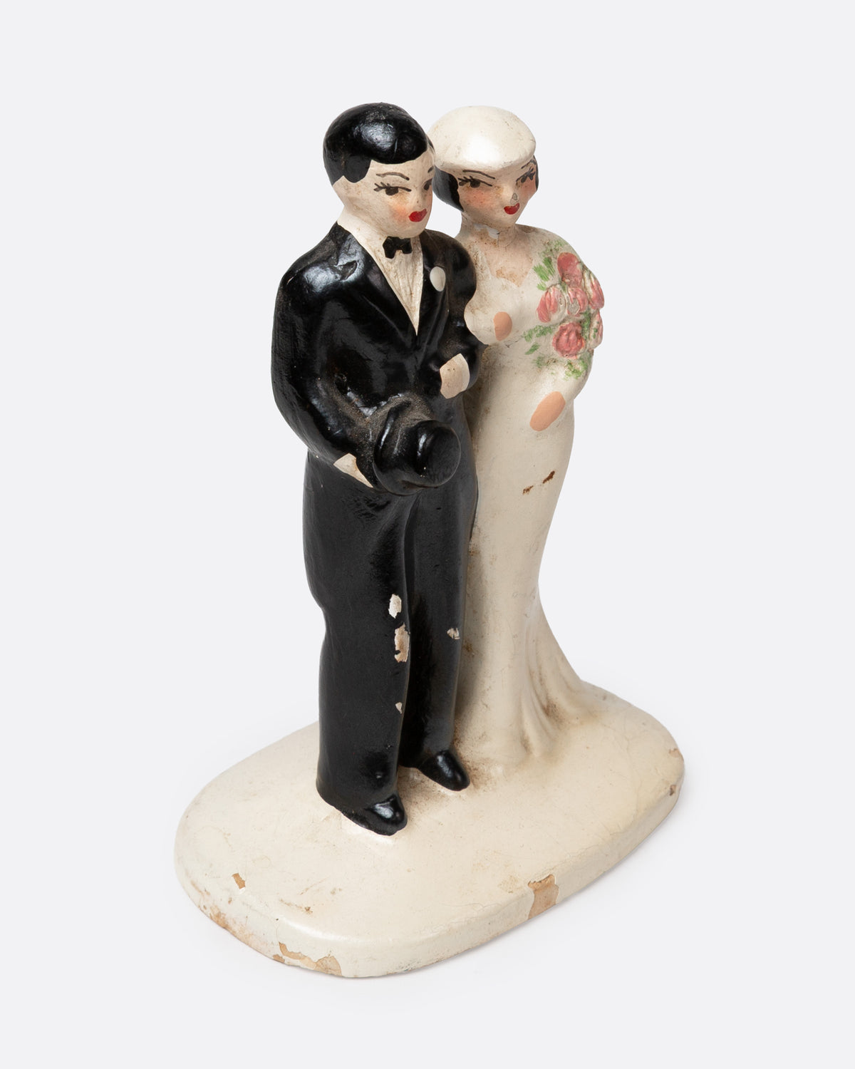 A ceramic wedding cake topper with a groom in a black tuxedo and a bride in a cream gown with pink floral details, shown from the side.