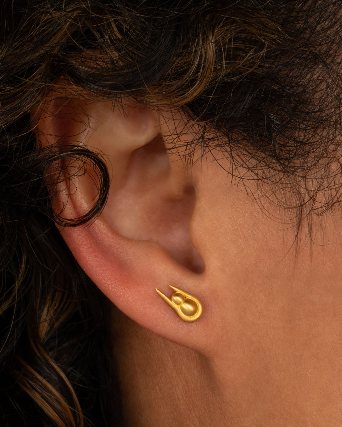 Lori Leven stud earring in matte yellow gold worn on an ear, a gold ball resting in a long gold horseshoe shape