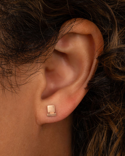 Lori Leven Square on a Bar stud earring in rose gold worn on an ear, a brushed gold square hanging below a short bar