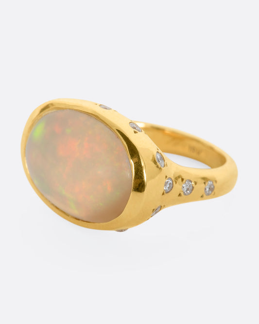 A polished yellow gold ring with a large east west oval Australian opal and diamonds on the setting and shoulders. View from the side.