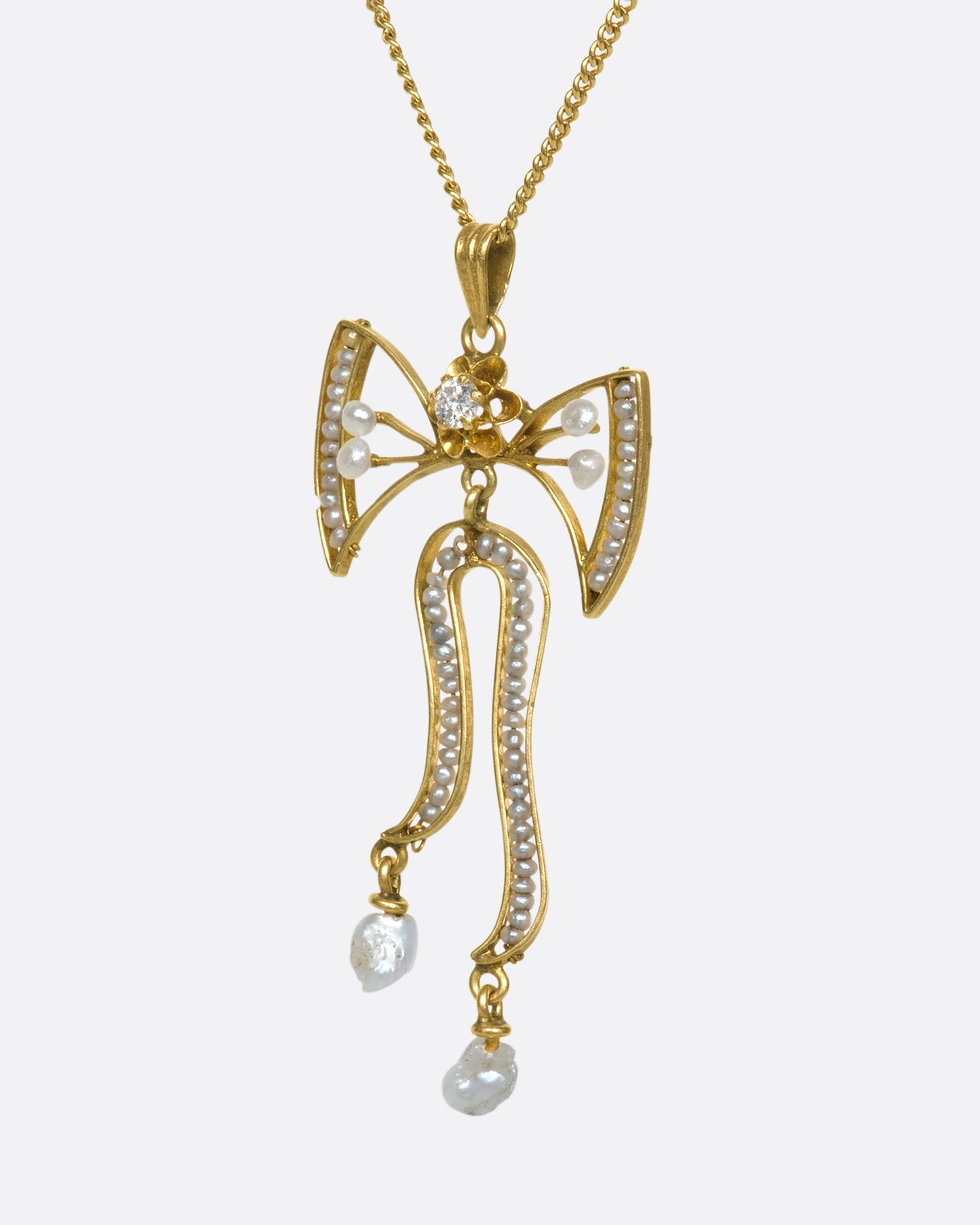 A slightly angled close up view of a yellow gold bow pendant with seed pearls and a diamond.