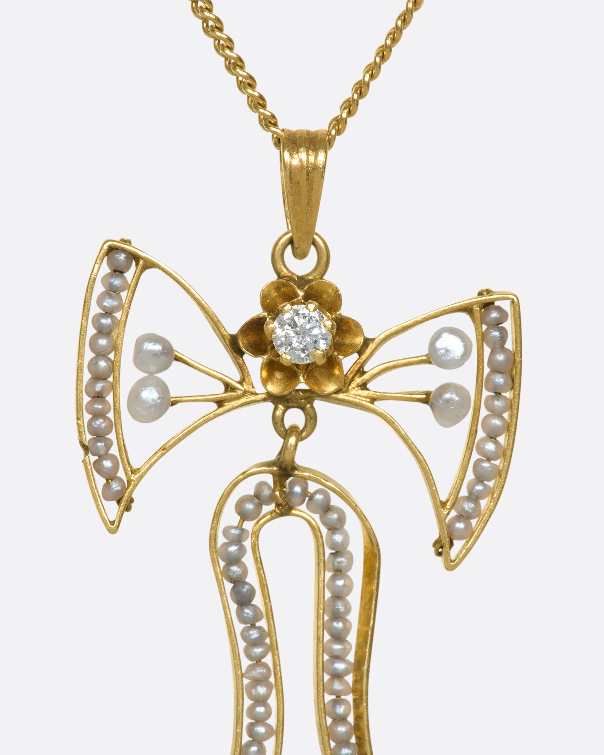 A close up view of a the top half of a yellow gold bow pendant with seed pearls and a diamond.