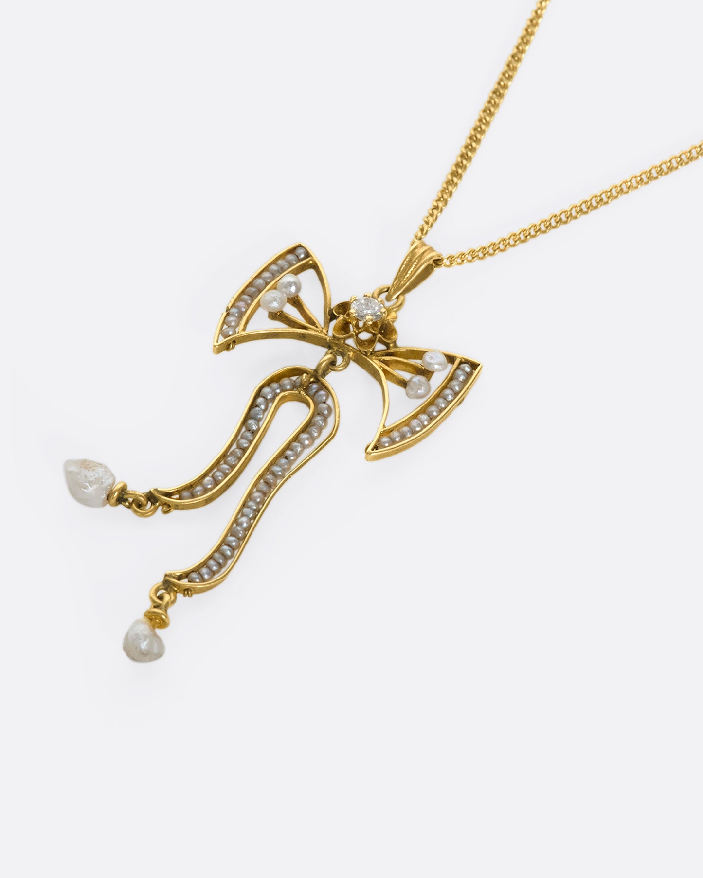 A slightly angled close up view of a yellow gold bow pendant with seed pearls and a diamond laying down flat..