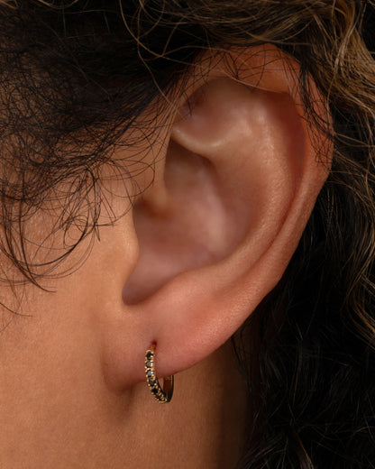 A yellow gold hinged hoop earring with black pavé diamonds, shown on a person.