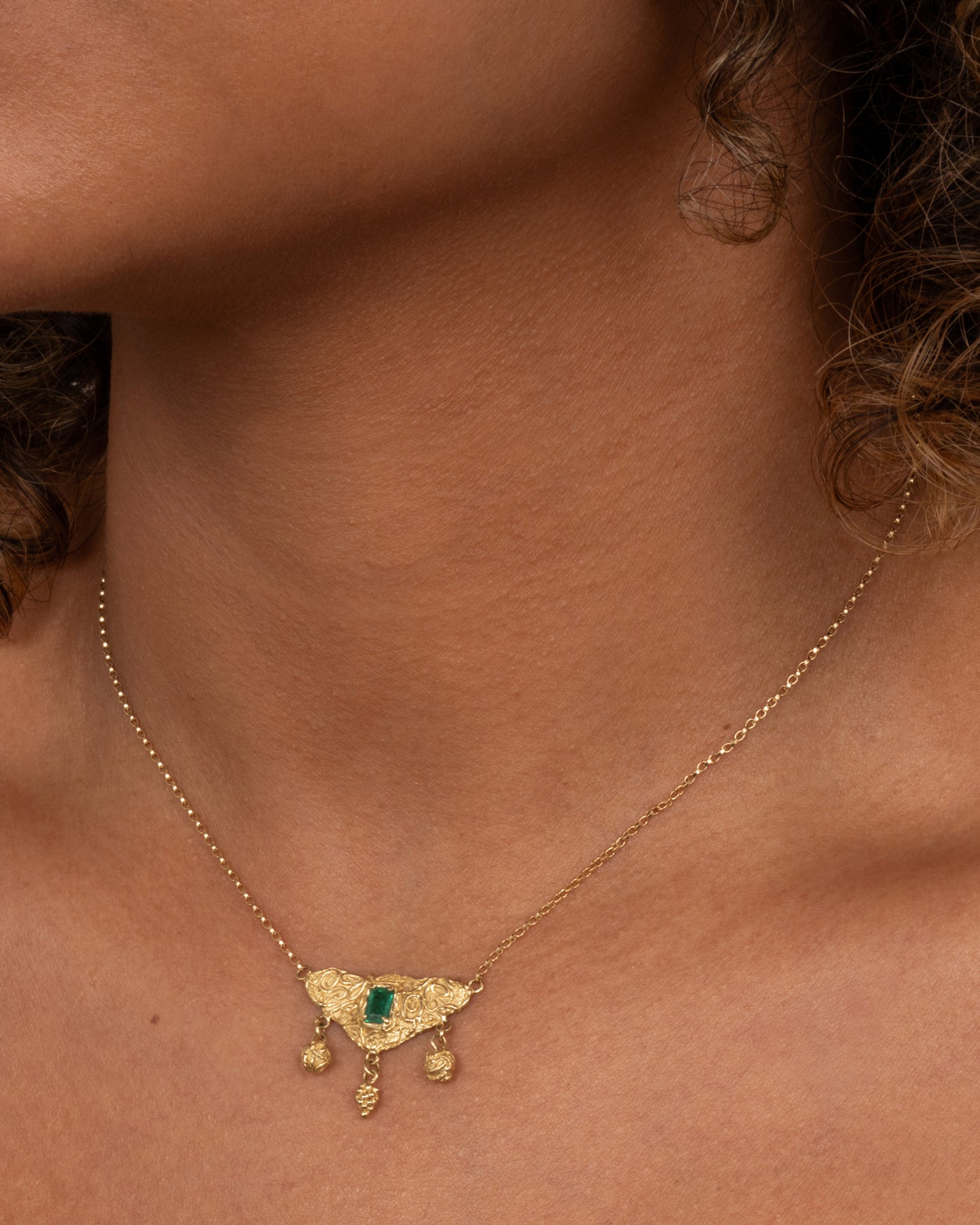 A yellow gold necklace with an emerald at the center of a sculptural pendant and three dangles- two blooming roses and a cluster of grapes. Shown worn around the neck.