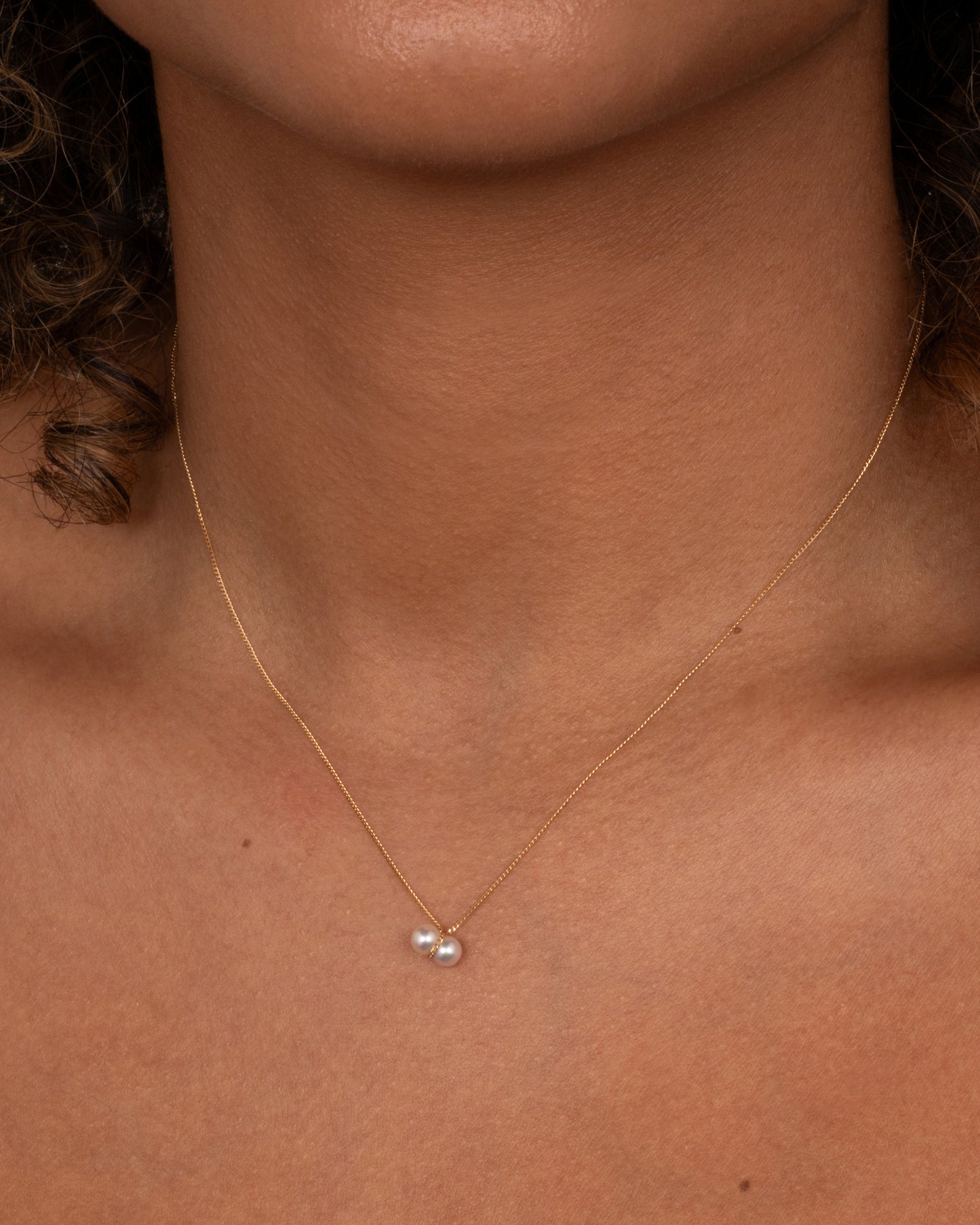 A yellow gold necklace with a small double pearl pendant. Shown worn around the neck.