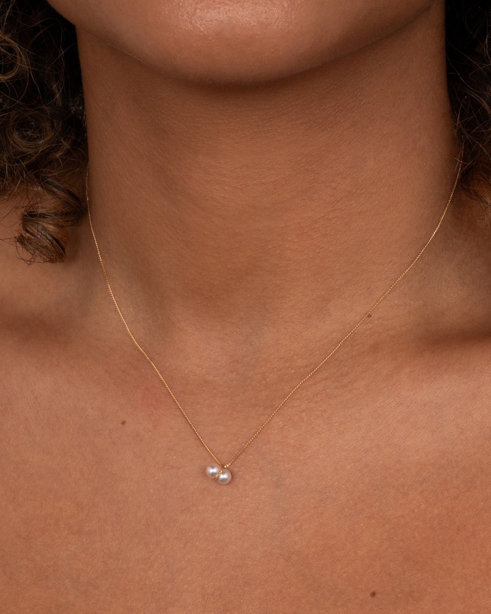 A yellow gold necklace with a small double pearl pendant. Shown worn around the neck.