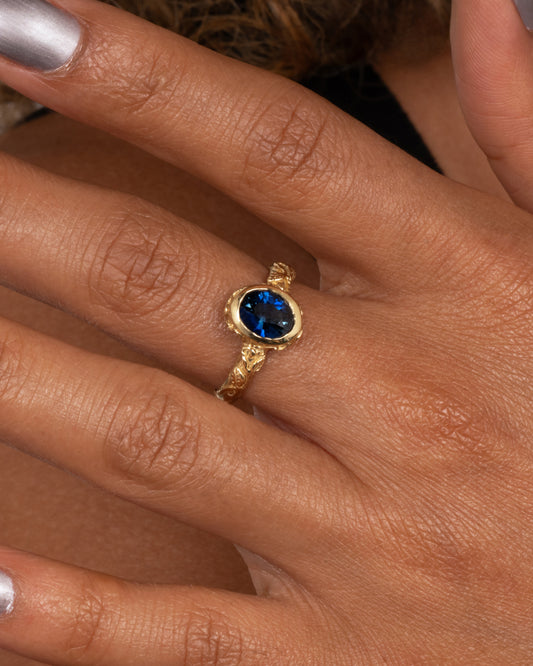 A yellow gold textured band with a teal blue sapphire. Shown worn on hand.