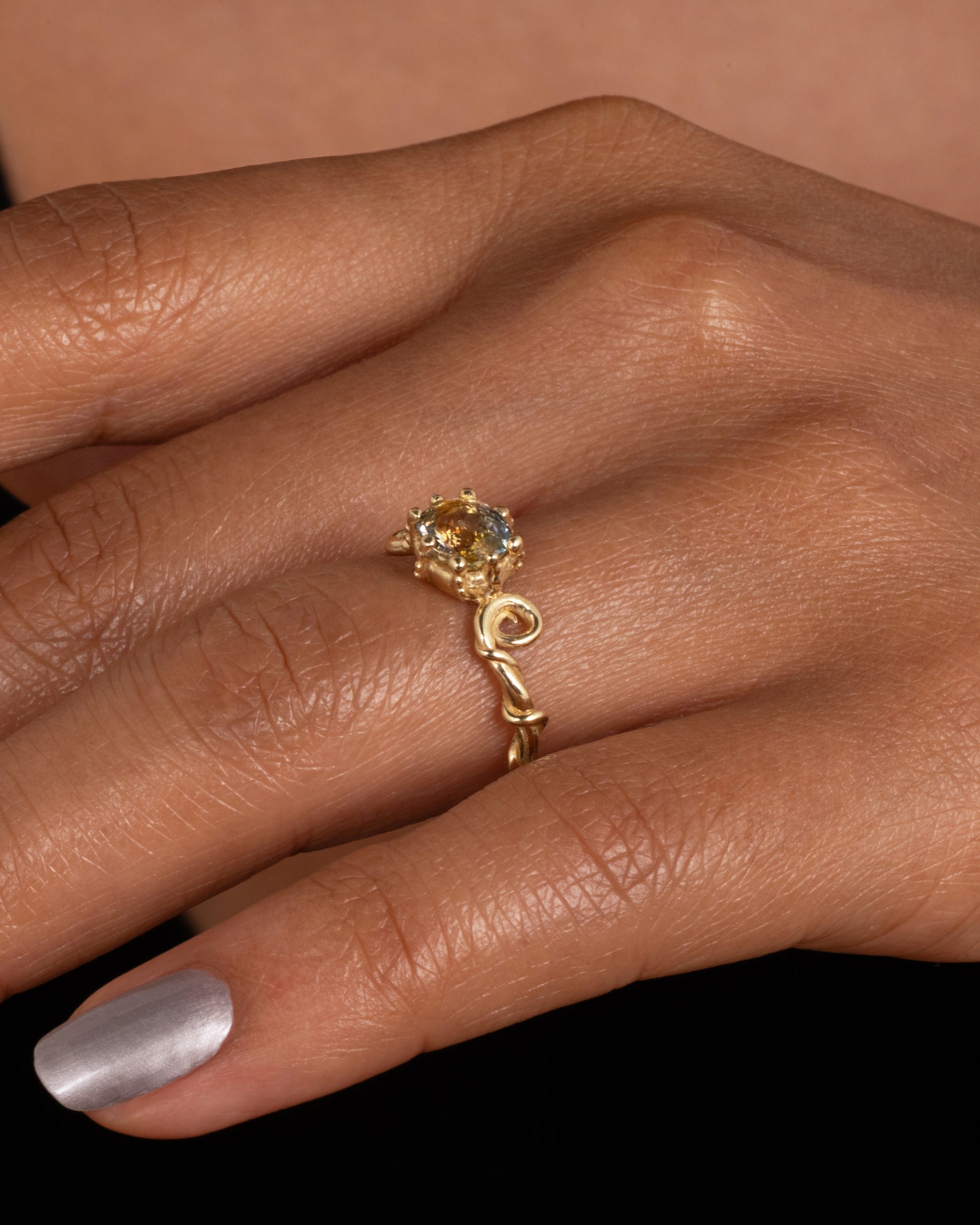 A twisty yellow gold ring with a bicolor sapphire, shown worn on finger.