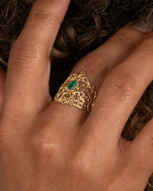 A wide yellow gold curling vine ring with  a central emerald. Shown worn on finger.