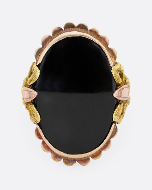 A yellow, rose, and green gold ring with a scalloped bezel, bow- topped shoulders, and an oval onyx. Shown from the front.