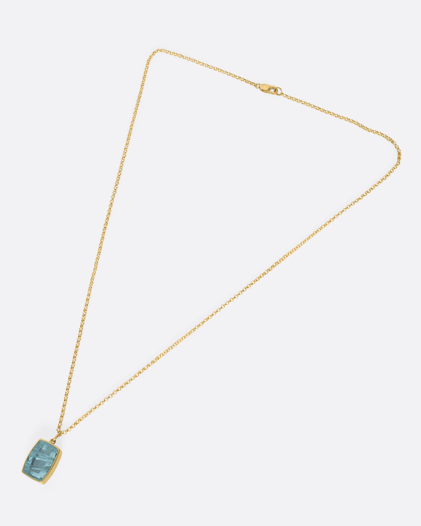 A cushion shaped aquamarine pendant with a yellow gold bezel on a gold chain laid down flat.