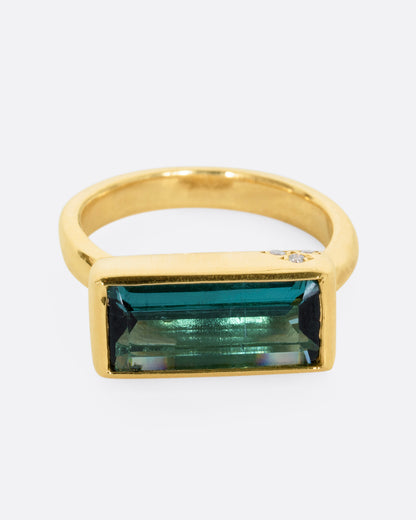 A yellow gold ring with a baguette indicolite tourmaline and small white diamonds. Shown from the front.
