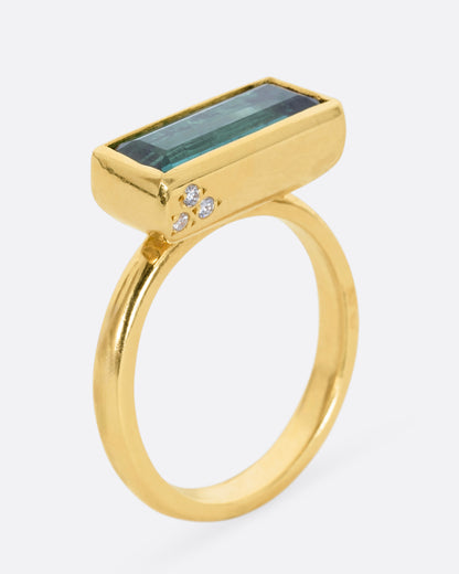 A yellow gold ring with a baguette indicolite tourmaline and small white diamonds. Shown from the side, standing up.