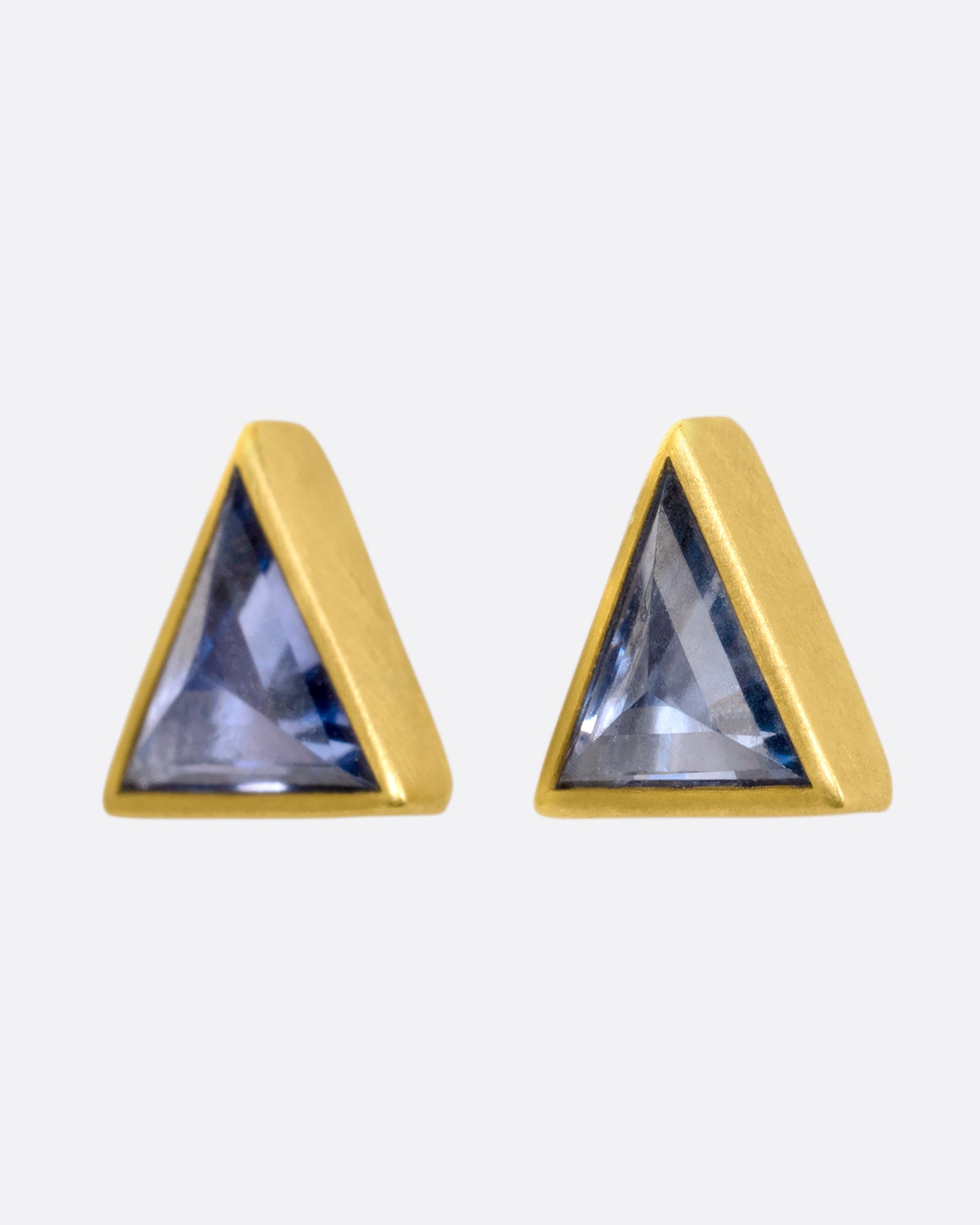 A pair of yellow gold triangle studs with pale blue bezel set sapphires. View from the side.