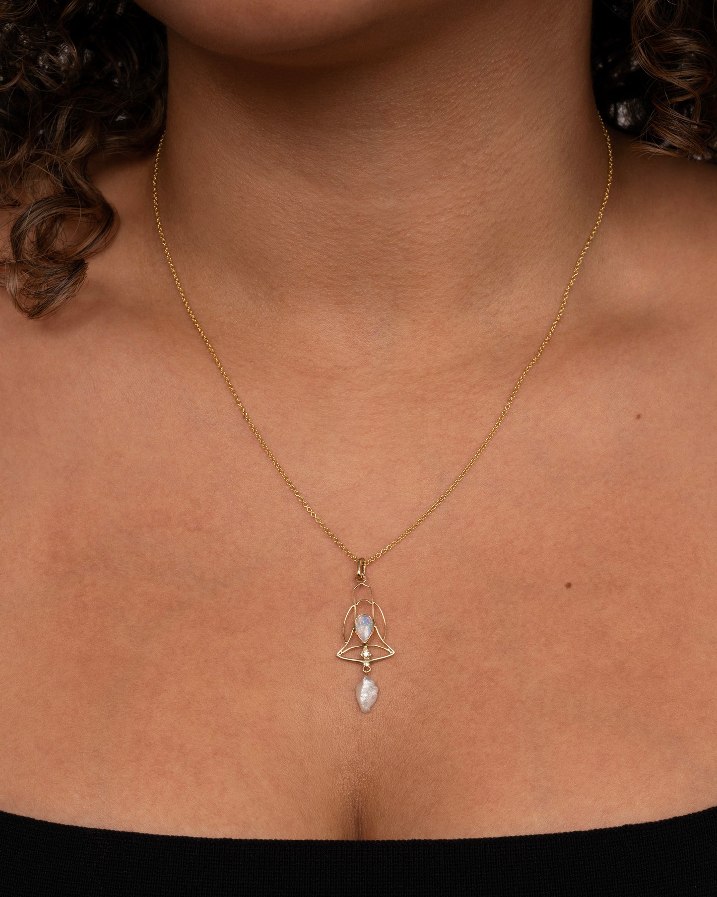 A yellow gold openwork bell pendant with an opal, diamond and dangling pearl. Shown worn around a neck.