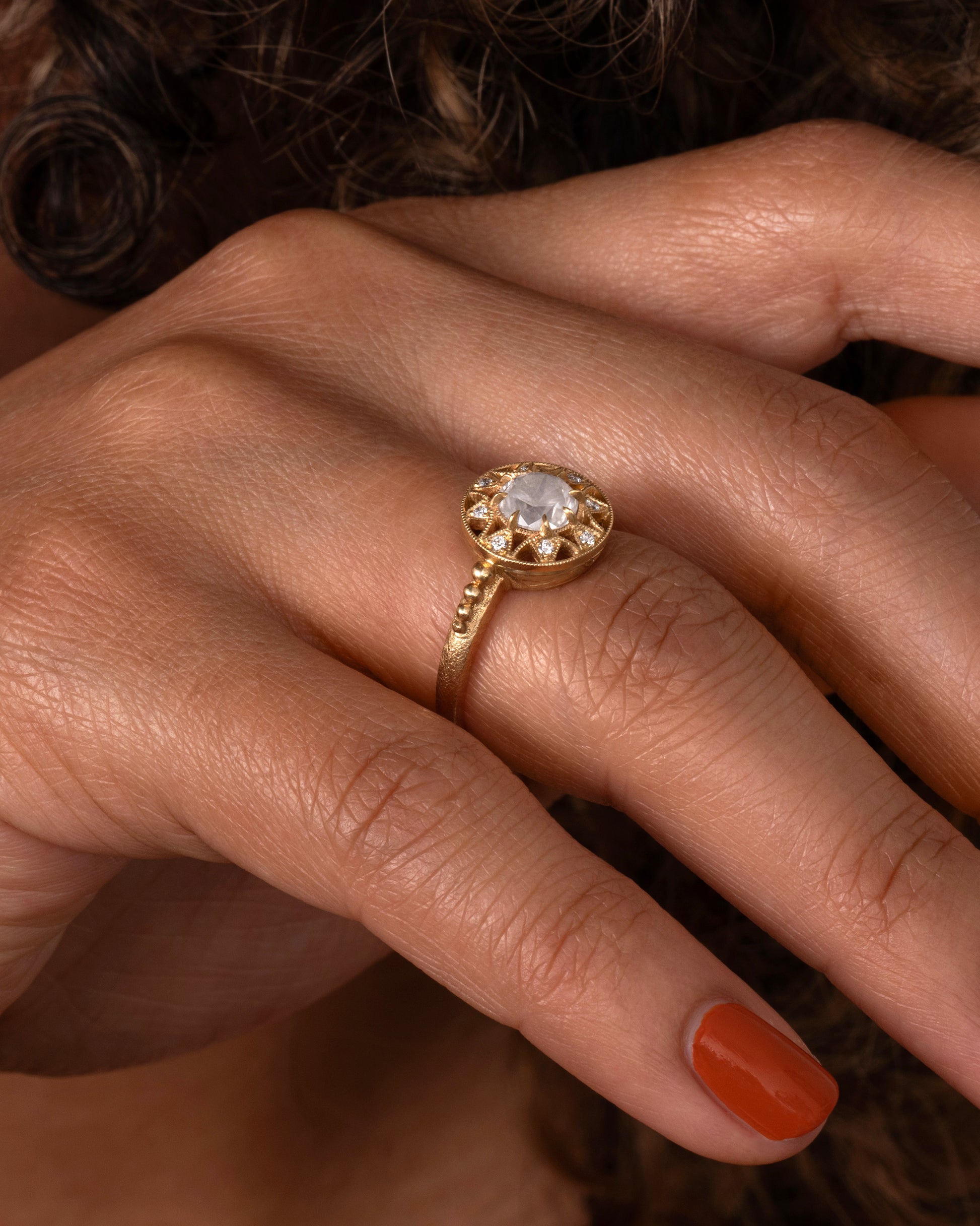 A yellow gold ring with a rose cut opalescent diamond at the center and a ring of brilliant cut diamonds around the face. Shown on a hand.