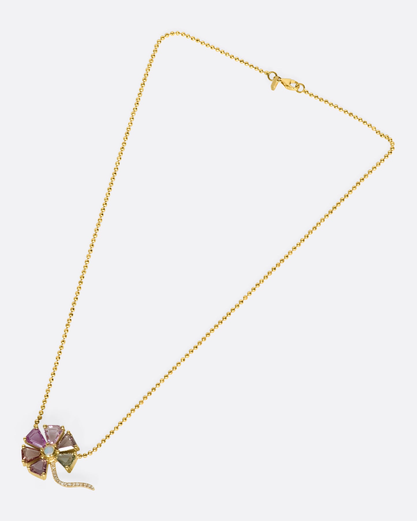 An overhead view of a flower pendant with sapphire petals, diamond stem, and opal center on a gold ball chain laying down flat.