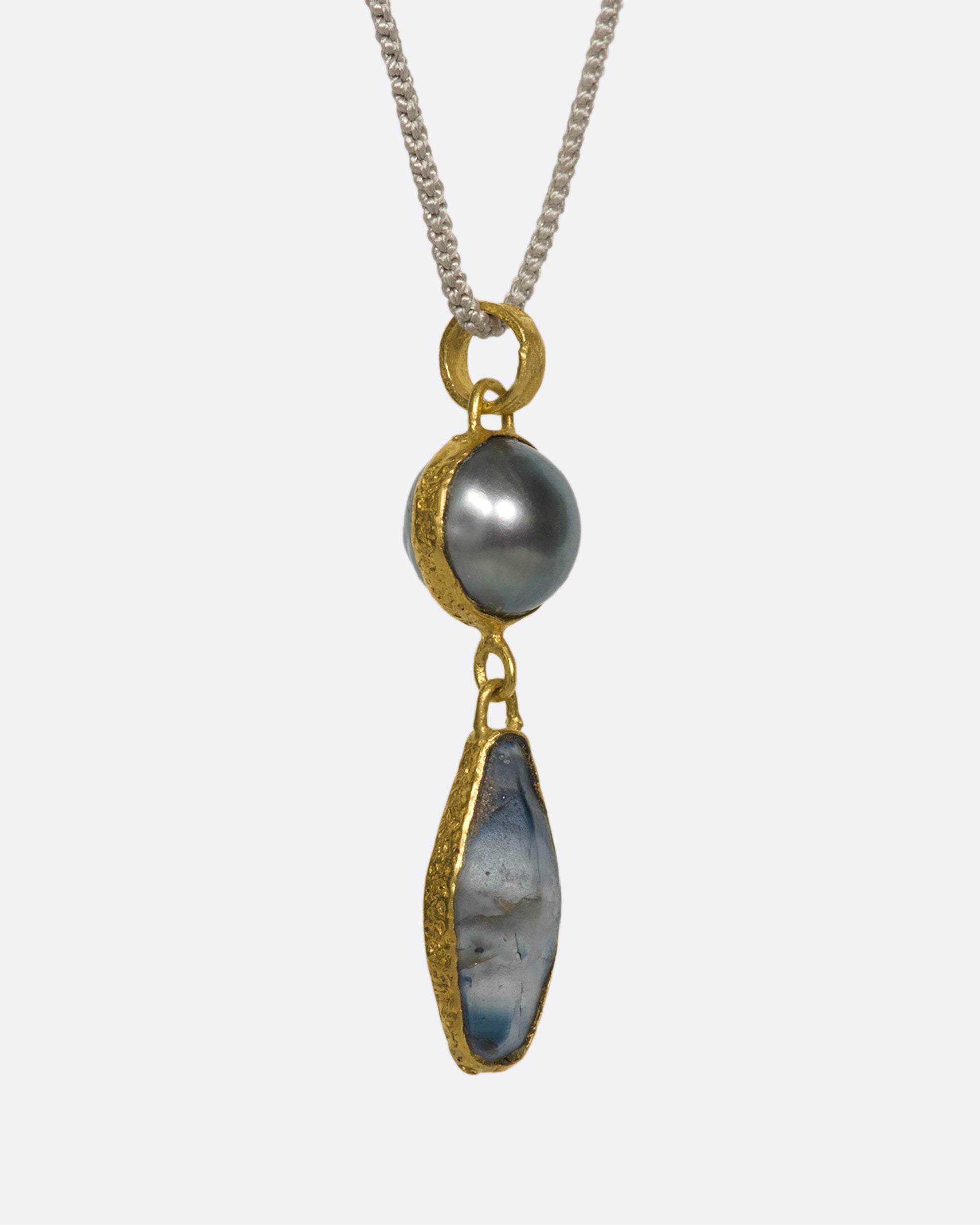 A close up of a hanging yellow gold pendant with a pearl and Sri Lankan sapphire on a cord.