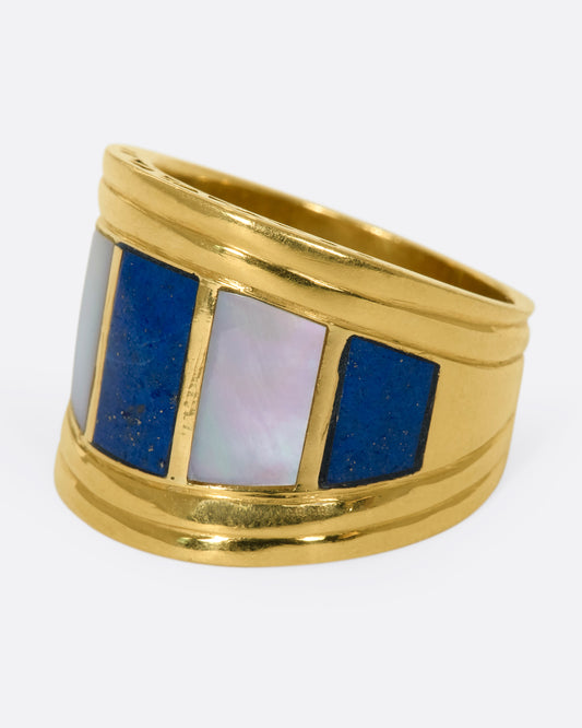 A left side view of a wide, tapered yellow gold ring with mother of pearl and lapis inlaid stripes.