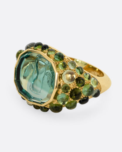 A yellow gold signet band with a hand carved winged hourglass in a bicolor green tourmaline. An array of green sapphire and tourmaline cabochons cover the band. Shown from the side.