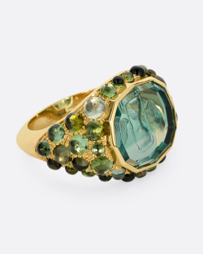 A yellow gold signet band with a hand carved winged hourglass in a bicolor green tourmaline. An array of green sapphire and tourmaline cabochons cover the band. Shown from the side.