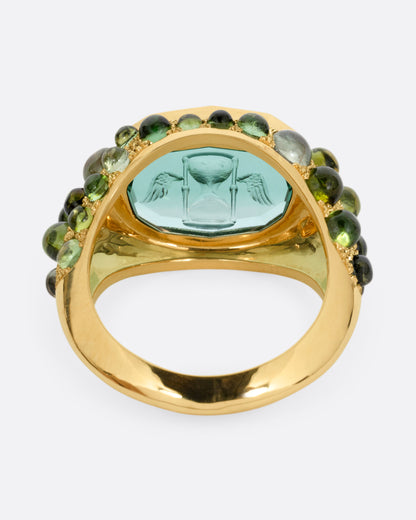 A yellow gold signet band with a hand carved winged hourglass in a bicolor green tourmaline. An array of green sapphire and tourmaline cabochons cover the band. Shown from the back.