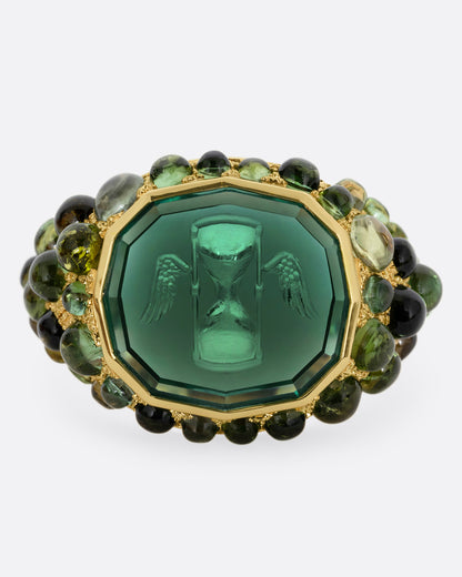 A yellow gold signet band with a hand carved winged hourglass in a bicolor green tourmaline. An array of green sapphire and tourmaline cabochons cover the band. Shown from the front.