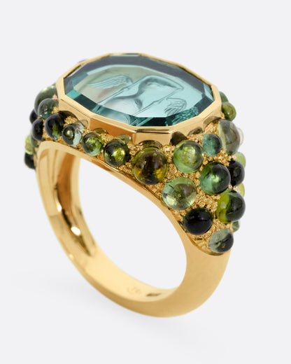 A yellow gold signet band with a hand carved winged hourglass in a bicolor green tourmaline. An array of green sapphire and tourmaline cabochons cover the band. Shown from the side standing up.