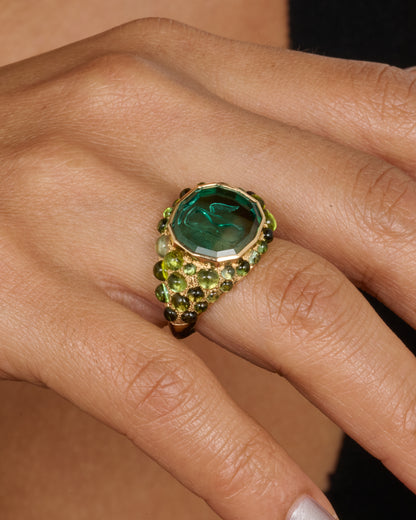 A yellow gold signet band with a hand carved winged hourglass in a bicolor green tourmaline. An array of green sapphire and tourmaline cabochons cover the band. Shown on a hand.