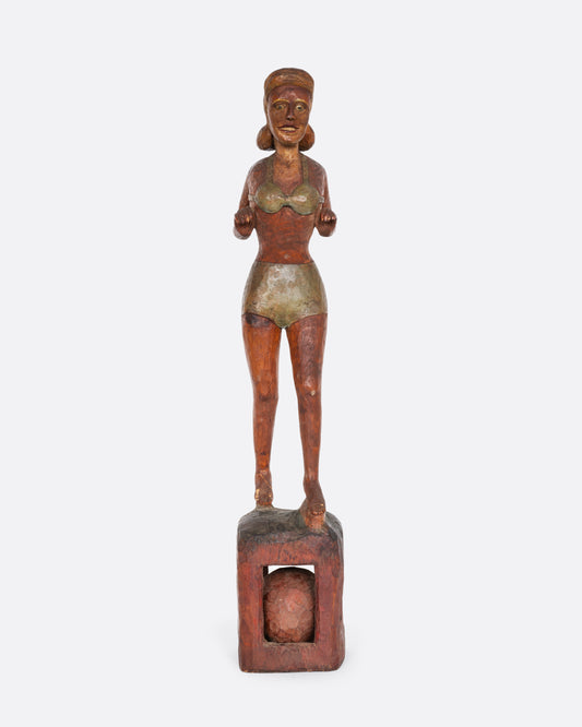 A hand carved wooden whimsy, a woman in a swimsuit standing on a ball. Shown from the front.