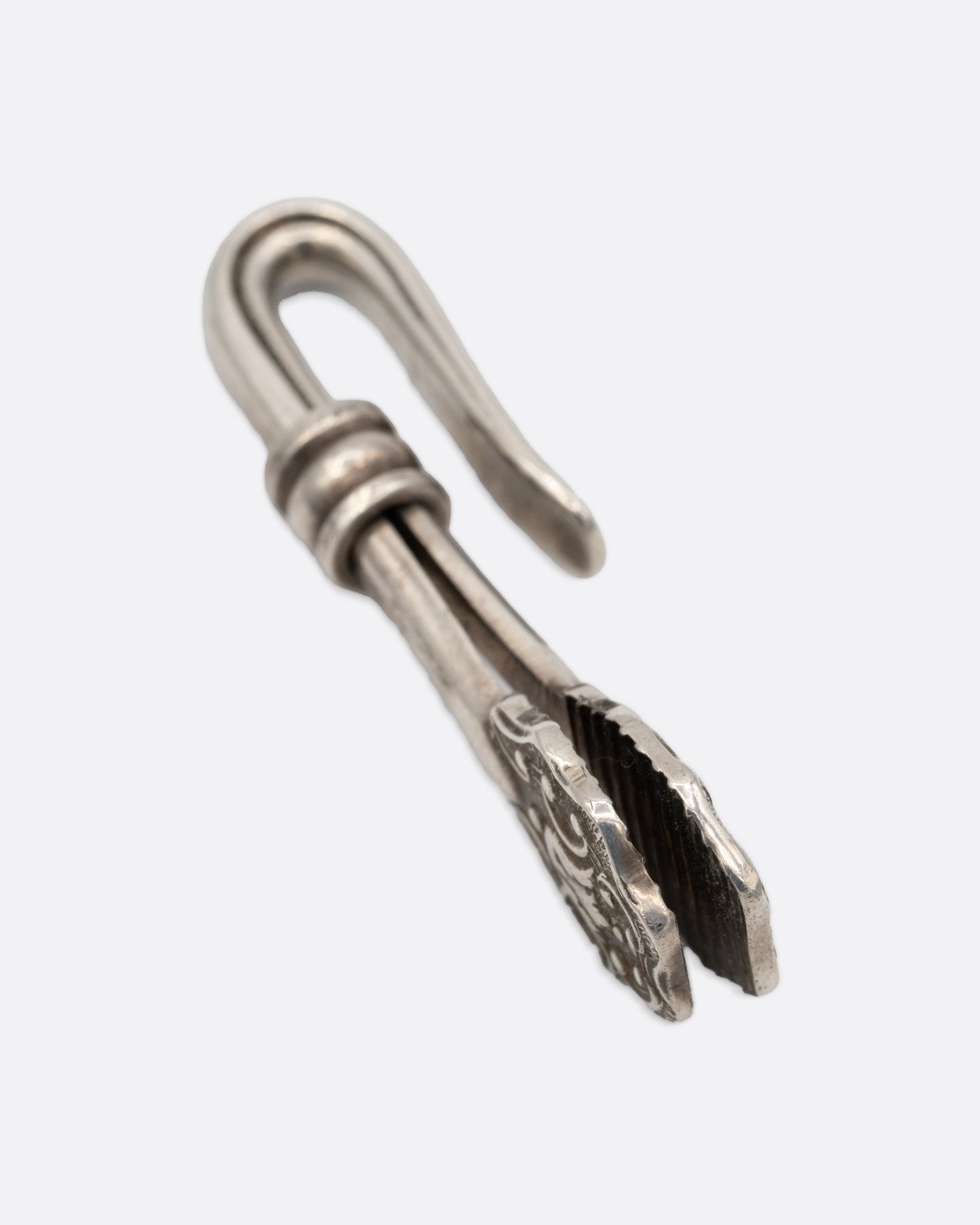 A silver engraved napkin holder with a  hook at one end and an oval clip to hold a napkin at the other. Shown from the side.