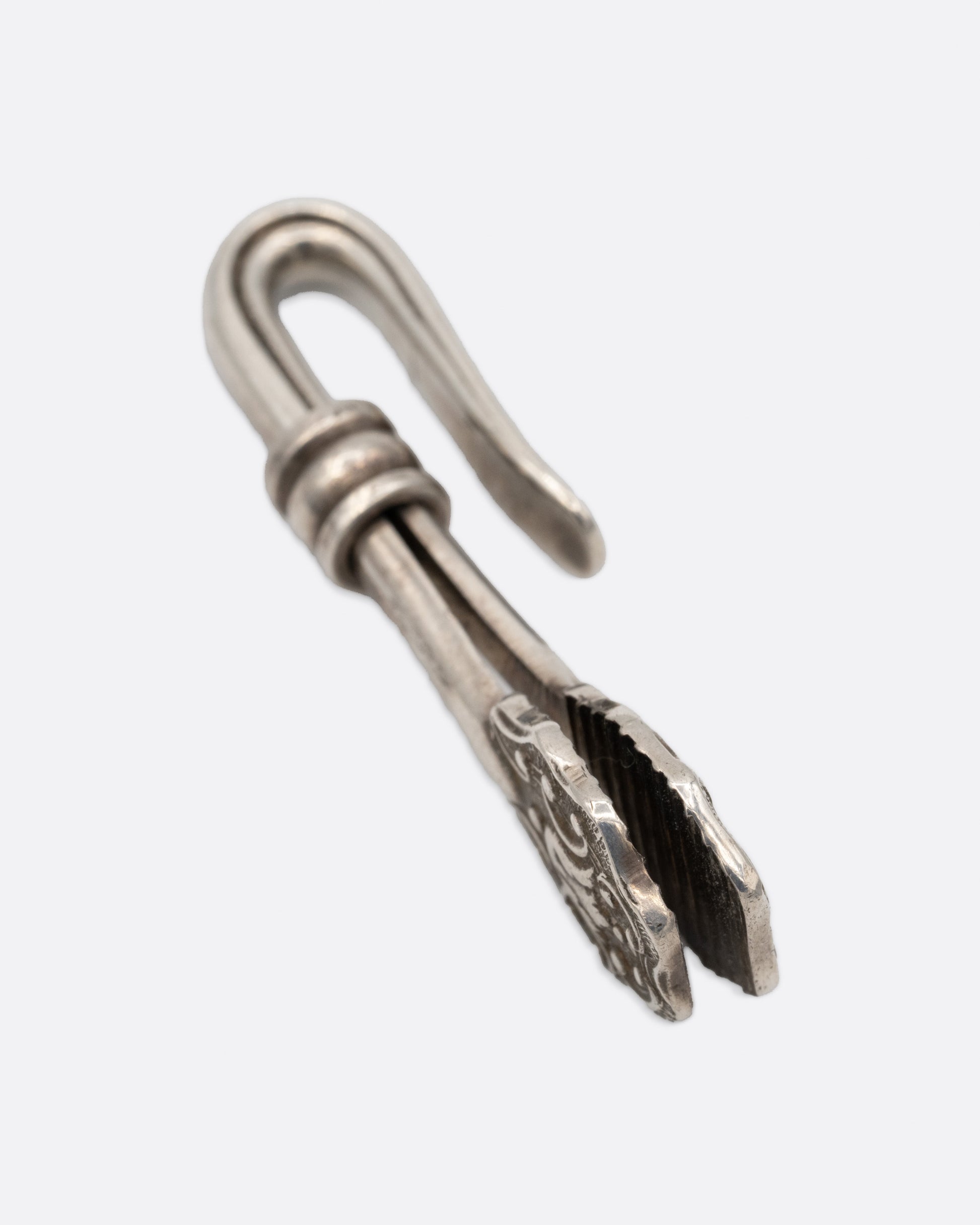 A silver engraved napkin holder with a  hook at one end and an oval clip to hold a napkin at the other. Shown from the side.