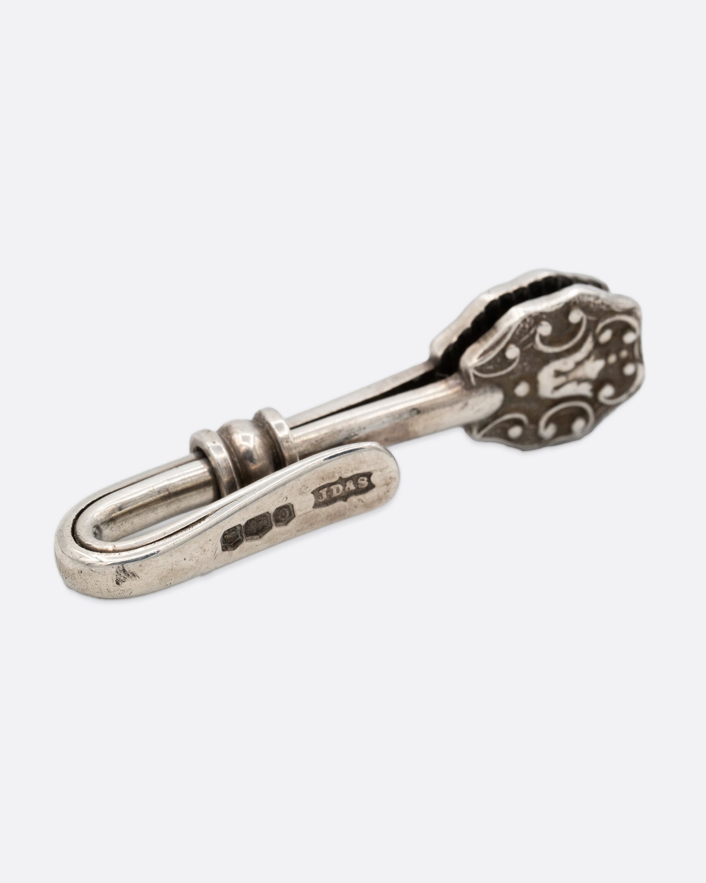 A silver engraved napkin holder with a  hook at one end and an oval clip to hold a napkin at the other. Shown from the back.