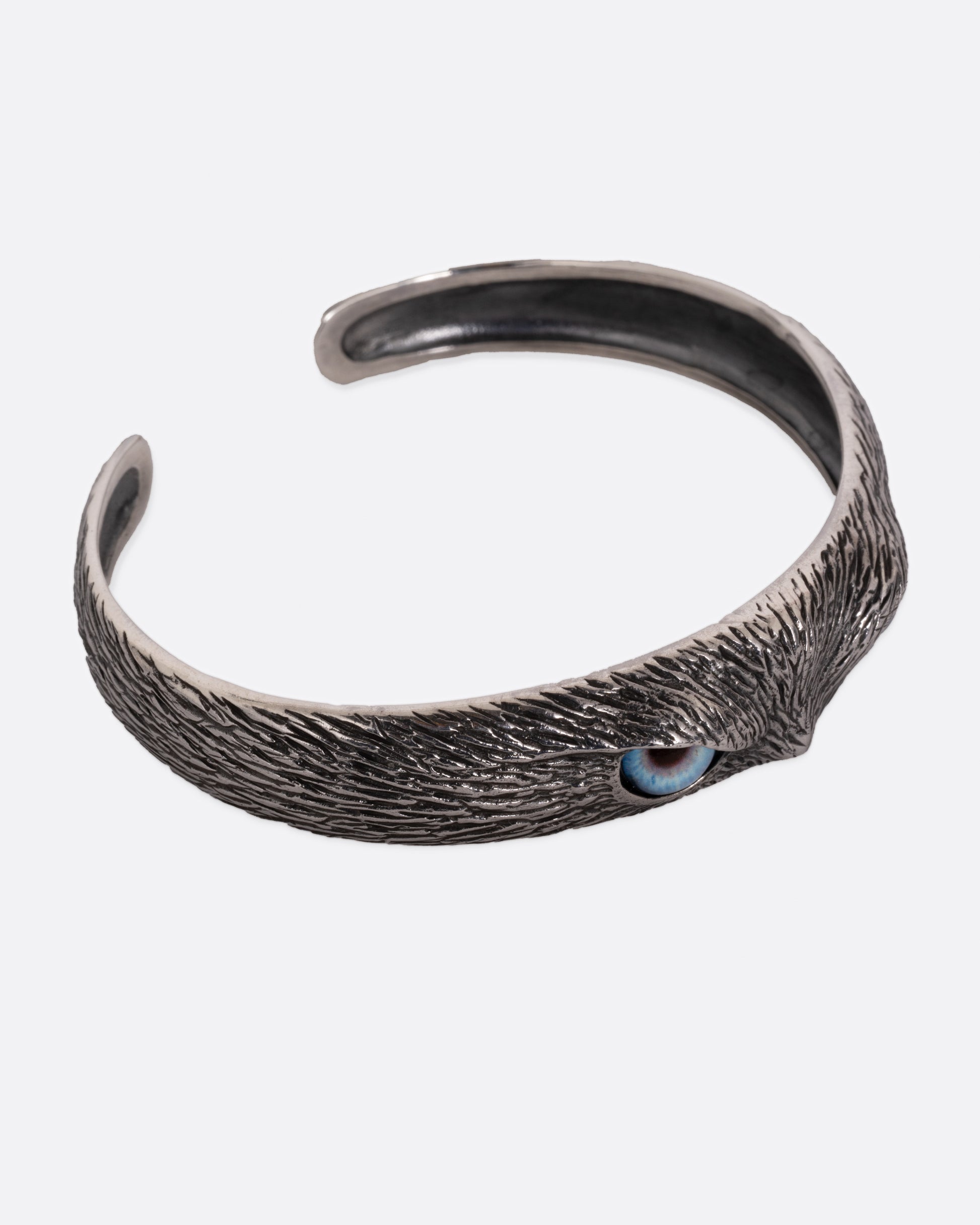 A silver bracelet with blue glass eyes and an all-over carved texture. Shown from the top on its side.