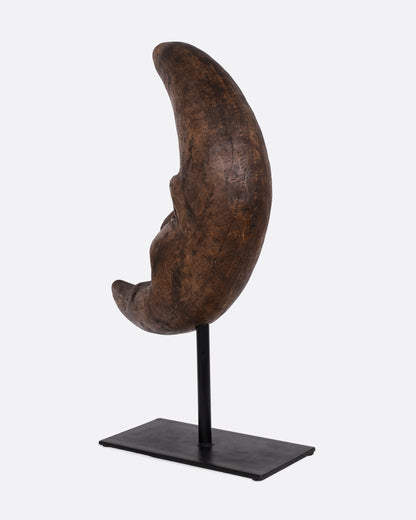 A wooden crescent moon takaan sculpture with a face on a black stand shown from the back.