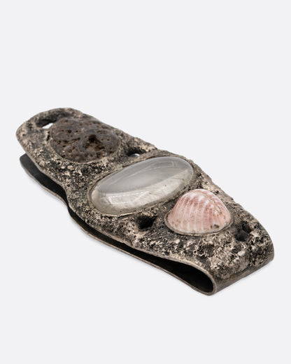A textural silver money clip with a  pebble, quartz, and seashell set in the front, shown from the side.