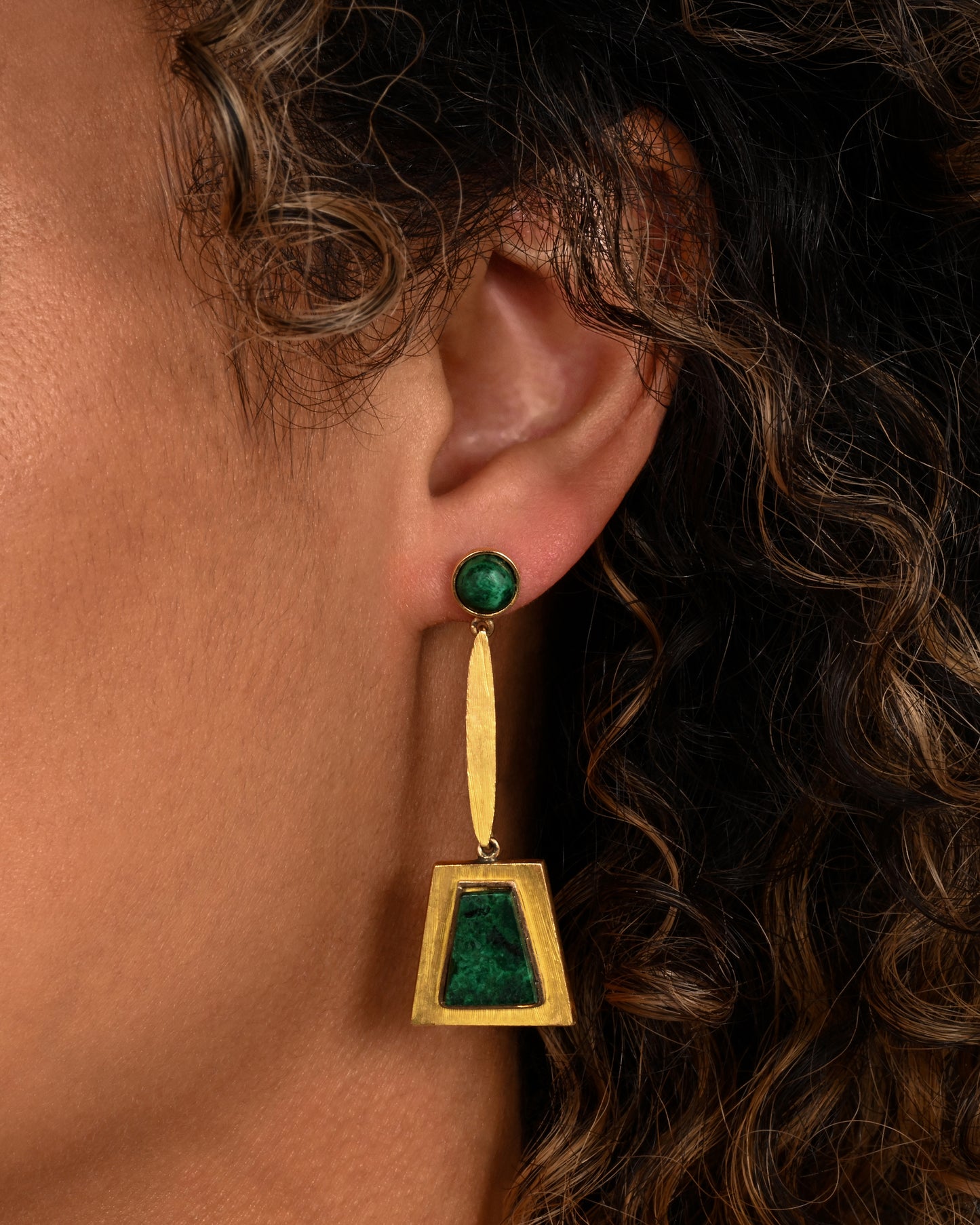 A yellow gold pair of trapezoid drop earrings with malachite. Shown on an ear.