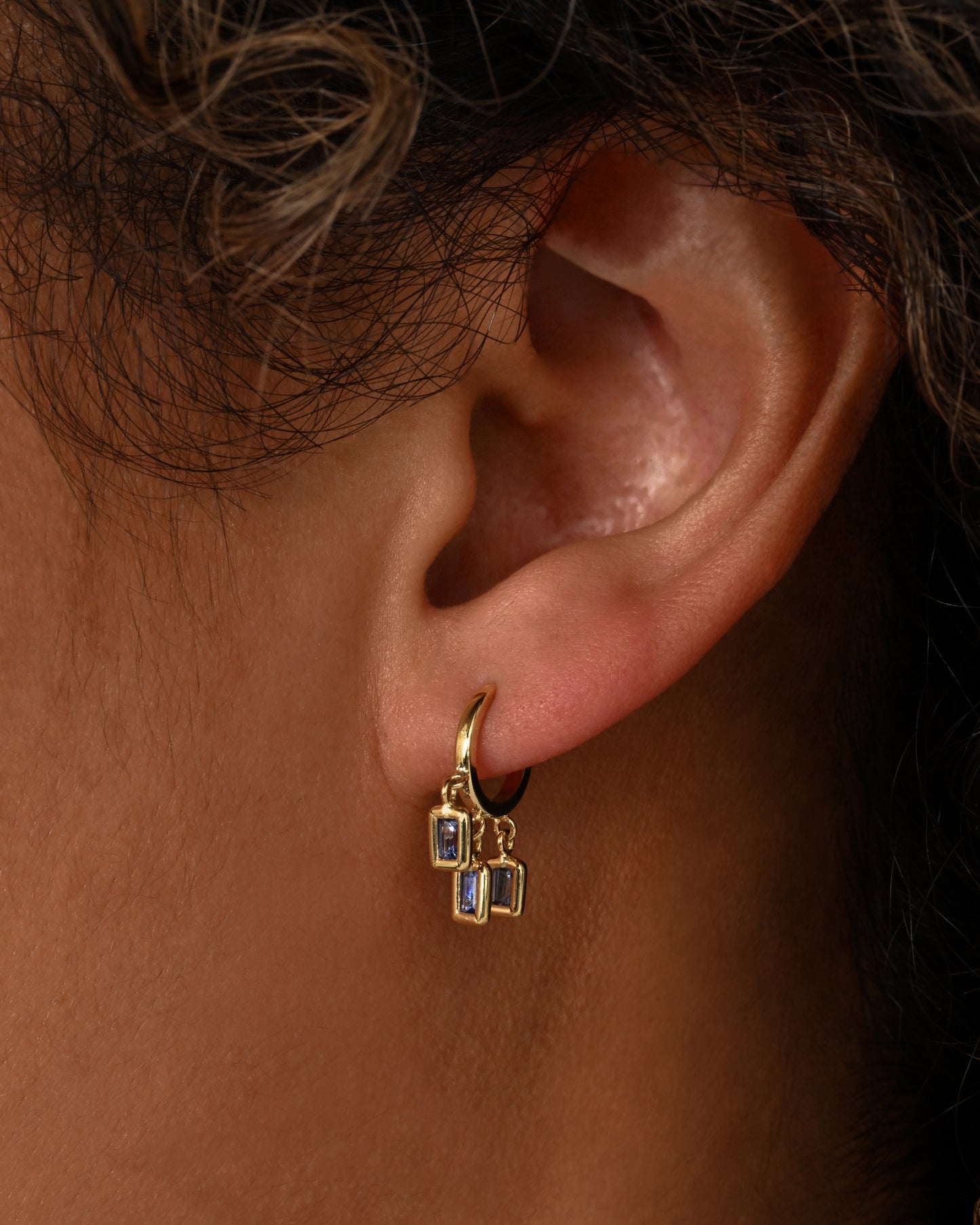 A yellow gold pair of hoop earrings with three dangling blue sapphires on each. Shown on an ear.