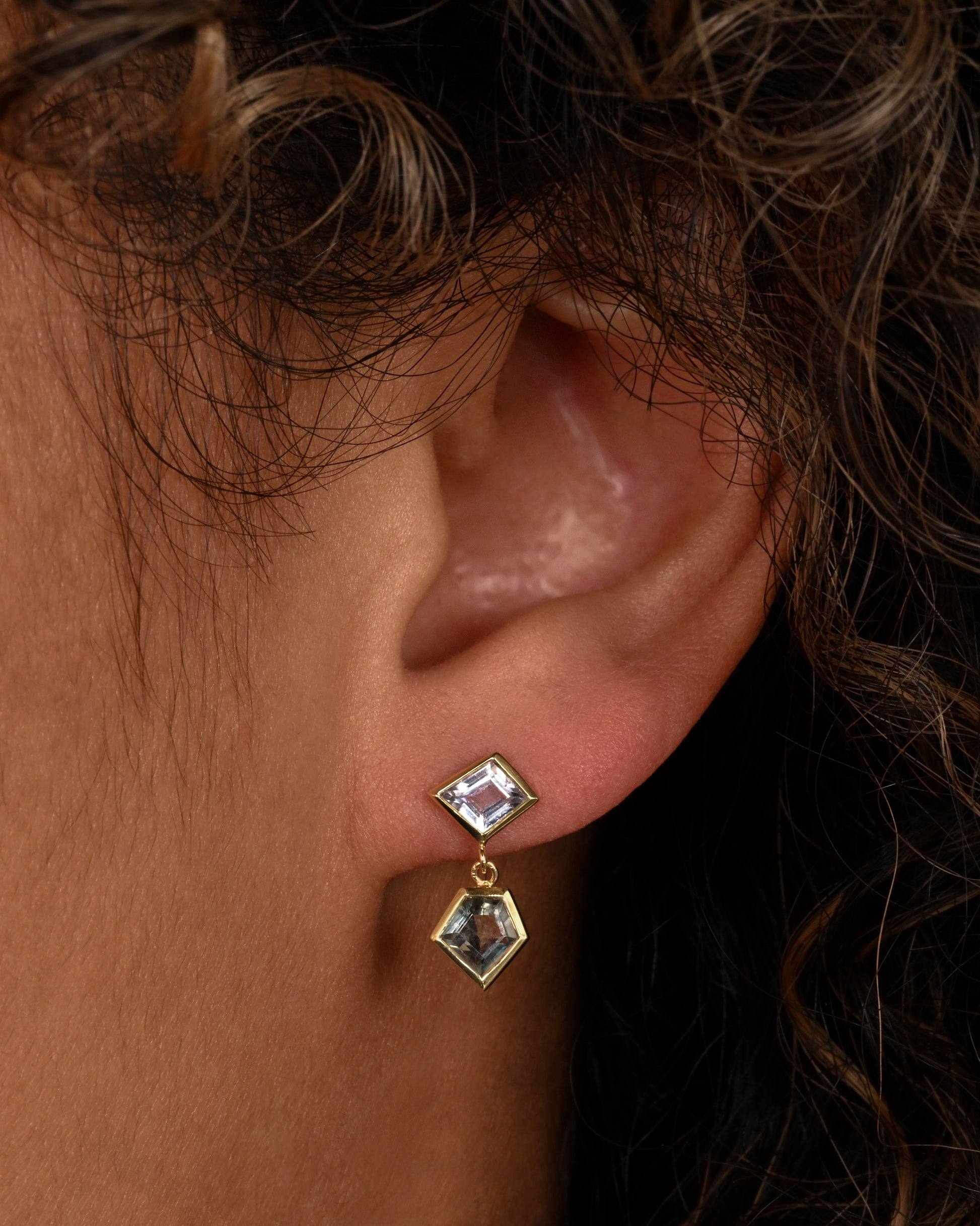 A pair of yellow gold drop earrings with trapezoid multicolor sapphires. Shown on an ear.